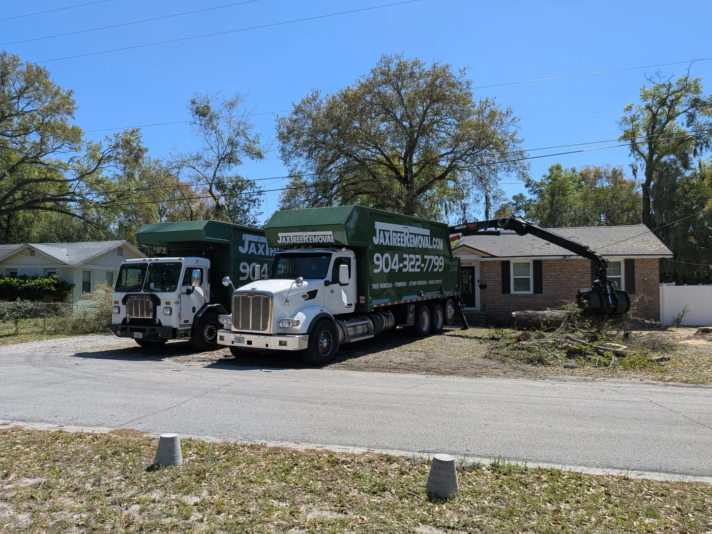 Two green Jax Tree Removal debris-hauling trucks with grapple loaders clearing brush and logs from a Jacksonville home after a storm